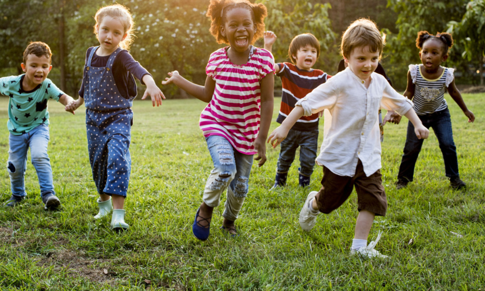 Childcare, early care, OST: Group young children holding hands while running towards camera across green grass with trees in background