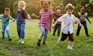 Childcare State Ballot Measures: Group young children holding hands while running towards camera across green grass with trees in background