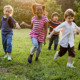 Childcare State Ballot Measures: Group young children holding hands while running towards camera across green grass with trees in background