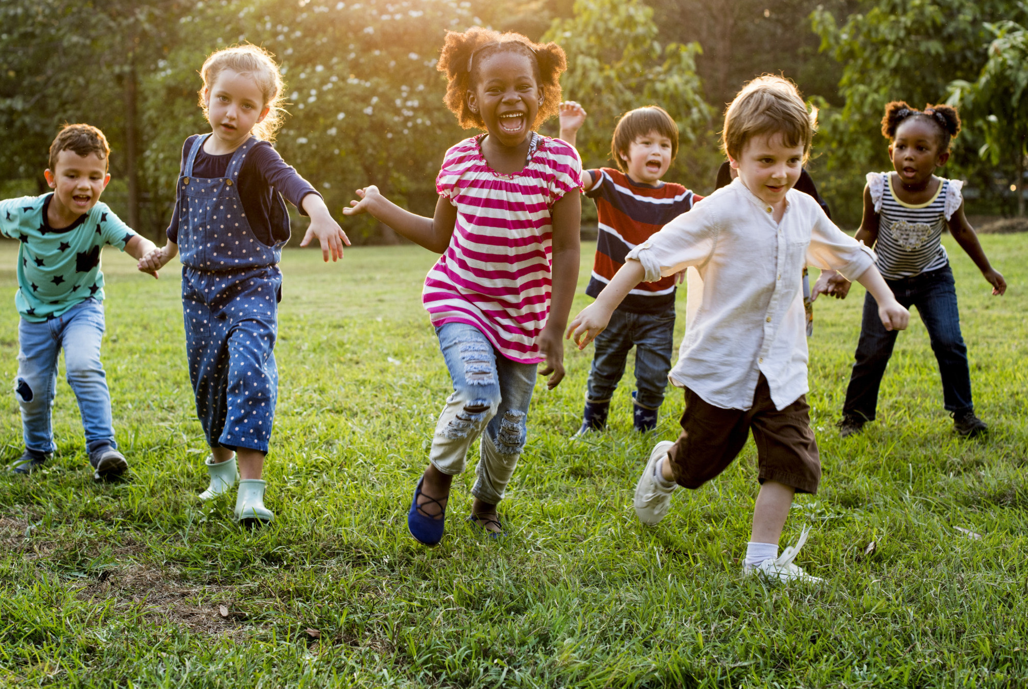 Childcare State Ballot Measures: Group young children holding hands while running towards camera across green grass with trees in background