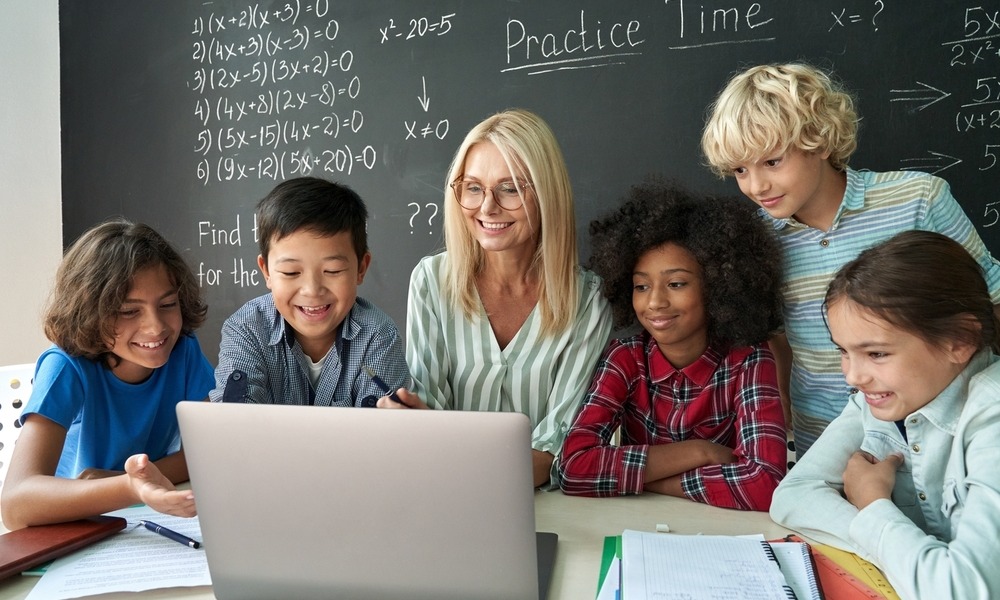 Teacher bias math students: Female teacher sits at her desk in front of blackboard with times tables and other math info on it, while several preteen students cluster around her looking at screen on her laptop