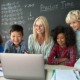 Teacher bias math students: Female teacher sits at her desk in front of blackboard with times tables and other math info on it, while several preteen students cluster around her looking at screen on her laptop