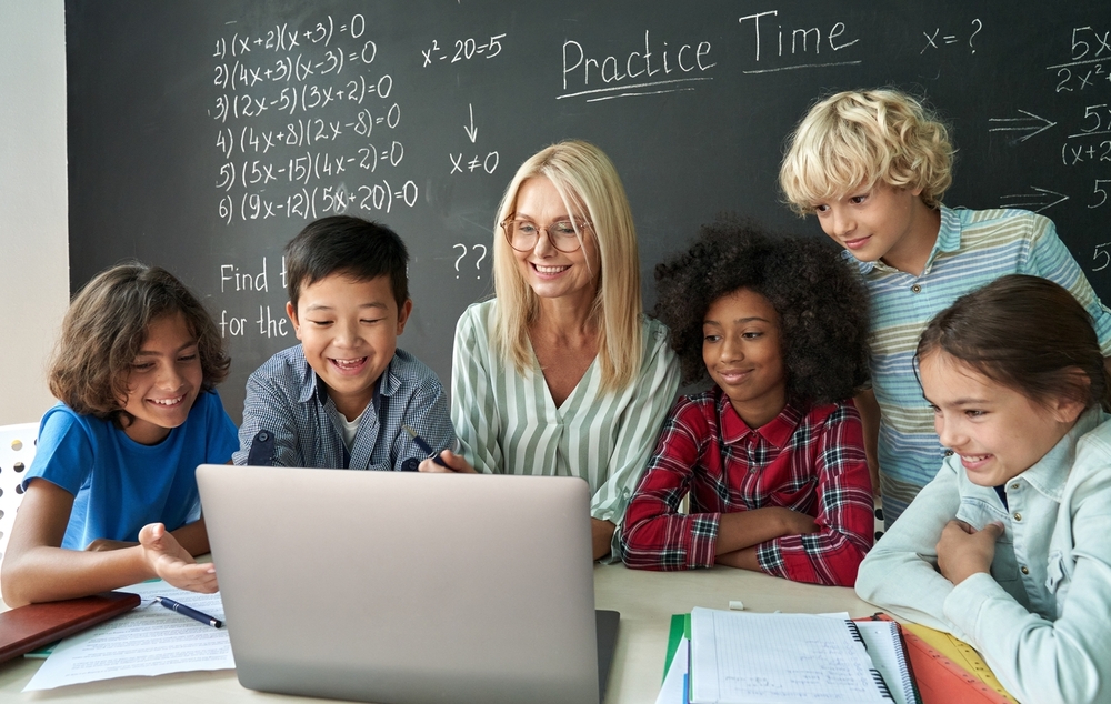 Teacher bias math students: Female teacher sits at her desk in front of blackboard with times tables and other math info on it, while several preteen students cluster around her looking at screen on her laptop