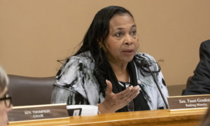 Foster care sleep-only placement: Black woman with long braids in black and white print jacket seated at table speaks into microphone gesturing with right hand