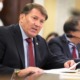 Shut down US DOE: Sebn. Mike Rounds, Reb. from S. Dakota -- a middle -aged man in navy suit and light blue shirt with red tie sits at dark wood table speaking into a microphone.