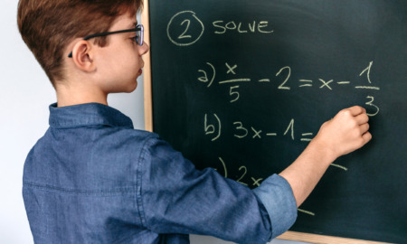 Algebra 8th grade options survey: Middle school age boy with short brown hair and glasses stands at chalkboard solving simple algebra equation