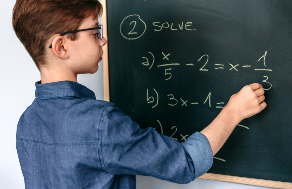 Algebra 8th grade options survey: Middle school age boy with short brown hair and glasses stands at chalkboard solving simple algebra equation