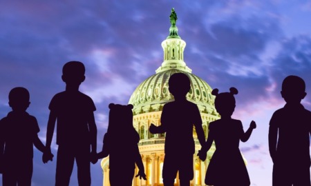 Child tax credit: Dome of US Capitol lit up against cloudy purple night sky with deep, dark purple silhouette of 6 children of various heights standing holding ands facing the Capitol overlaid in foreground.