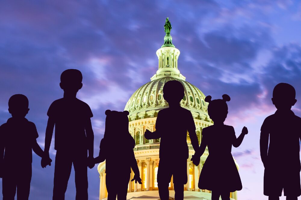 Child tax credit: Dome of US Capitol lit up against cloudy purple night sky with deep, dark purple silhouette of 6 children of various heights standing holding ands facing the Capitol overlaid in foreground.