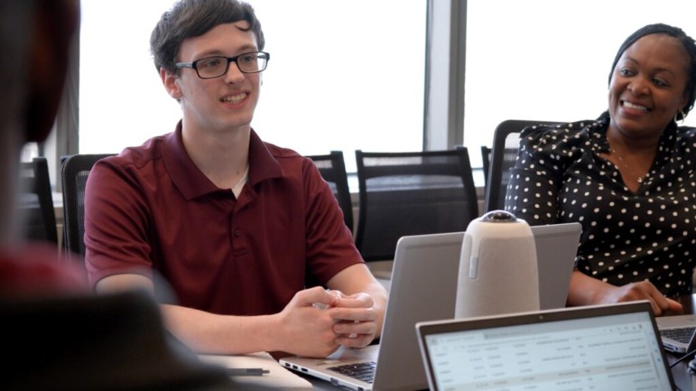 Apprenticeships Indiana: A young man in dark frame glsses and a burgundy polo shirt sits at desk in front of laptop with hands crossed next to a older Black woman in a black & white polka-dot dress speaking to others in the group seated at the table