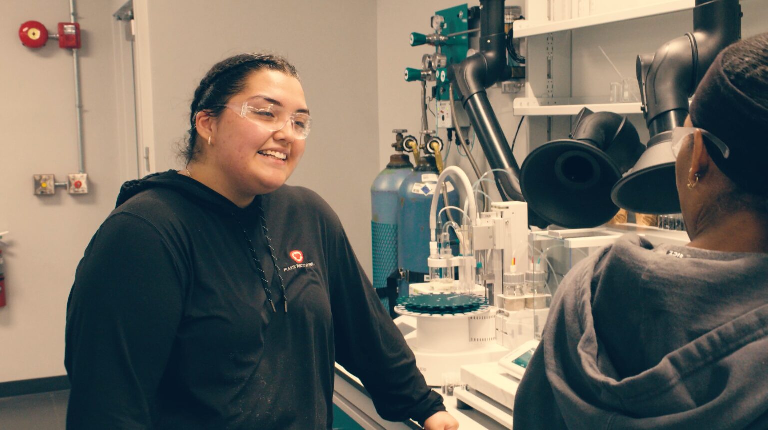 Apprenticeship Indiana: Young woman with dark hair pulled back wearing clear safety glasses and a navy sweatshirt, stands at work table covered with plastic recycling machinery speaking to another young adult.