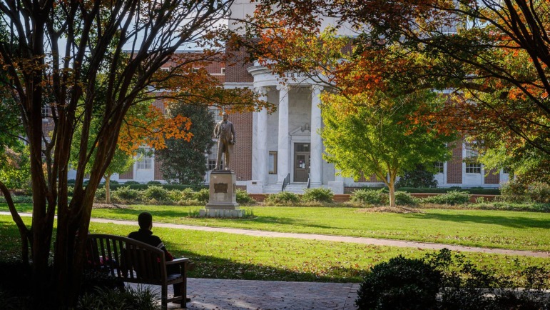 Rural college major elimination: Silhouette of person sitting on benchunder tall trees with fall colors looking across green lawn to traditional 3 story red brick building wit white pillars