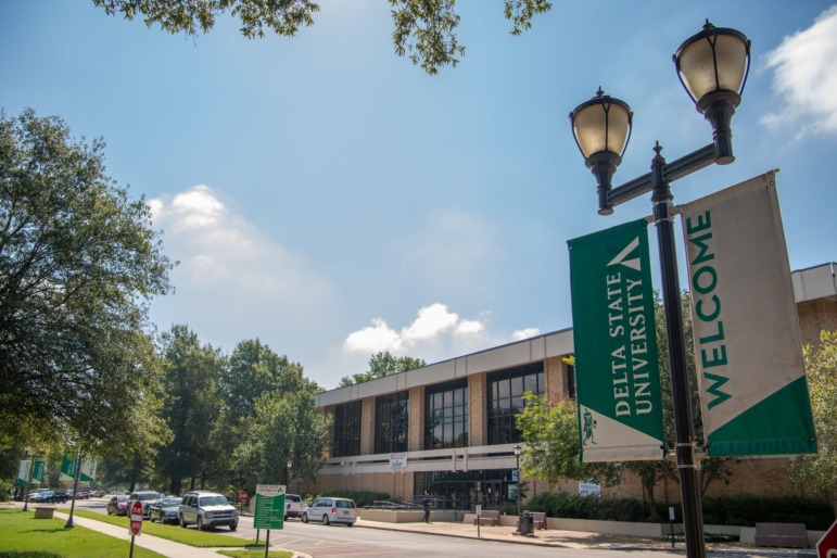 Rural colleges closing programs: Banner sign "Delta State University - Welcome" on black lamppost with multistory, modern, red brick building in background.