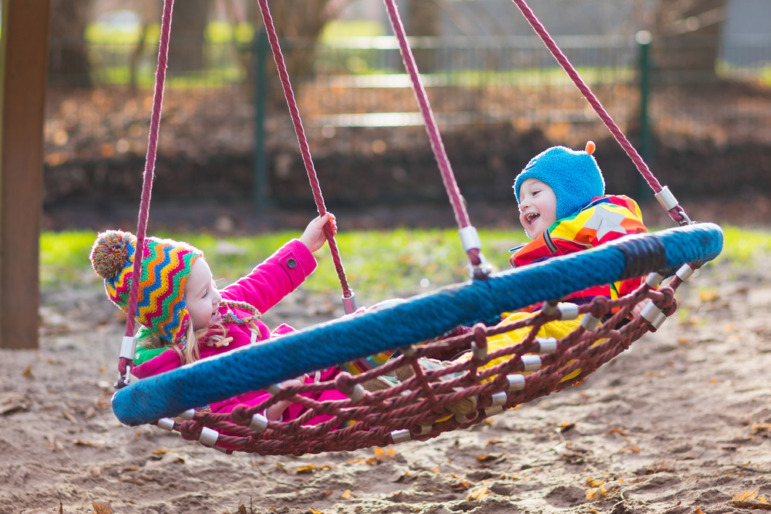 Childcare-Preschool dark future: Two toddlers in winter coats and hats sit on woven circle swing twirling around in sandy area of playground