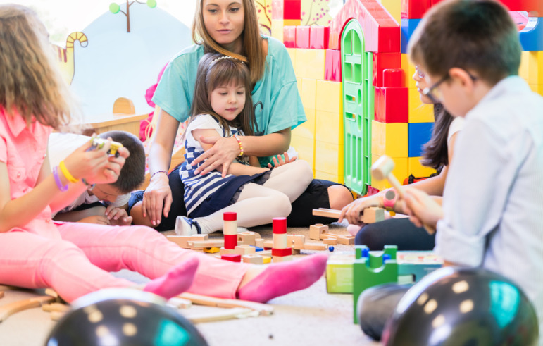 Childcare-Preschool dark future: Female teacher sits in circle on floor, with several toddlers with block toys in the center, holding one little girl in her lap, in preschool classroom