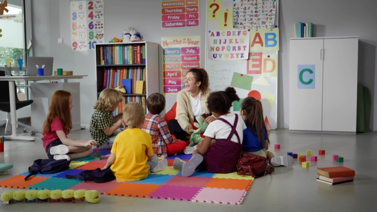 Childcare-Preschool dark future: Laughing female teacher sits on floor in a circle with several preschool students with colorful bulletin board and bookcase in background