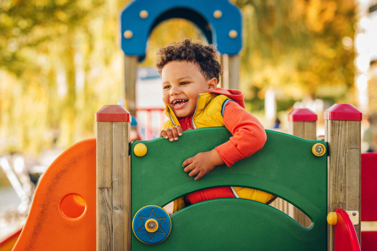 Childcare-Preschool dark future: Black boy toddler in winter jacket plays on colorful jungle gym in outdoor playground