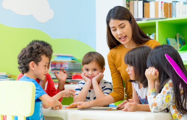 Childcare-Preschool dark future: Teen with long dark hair sits at preschool table talking to several toddler students 