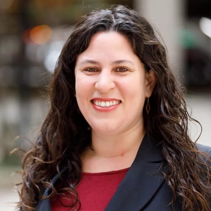 College listening cohort: Headshot woman with long brunette curly hair in dark suit with red top