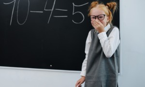 US Math scores decline: Young girl wit red hair in buns, black-framed glassses, wearing a gray and white dress, stands at blackboard holding chalk and giggling because she made a subtraction mistake: 10-4=5.