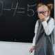 US Math scores decline: Young girl wit red hair in buns, black-framed glassses, wearing a gray and white dress, stands at blackboard holding chalk and giggling because she made a subtraction mistake: 10-4=5.