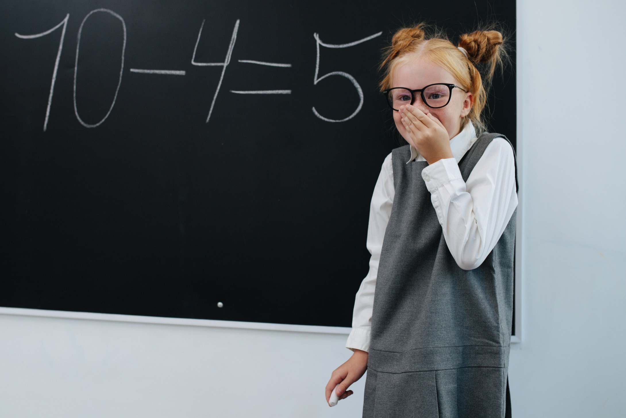 US Math scores decline: Young girl wit red hair in buns, black-framed glassses, wearing a gray and white dress, stands at blackboard holding chalk and giggling because she made a subtraction mistake: 10-4=5.