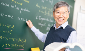 High school calculus survey: Older Asian male with gray hair and glasses in white business shirt with navy vest stands at chalkboard writing calculus formulas while smiling towards students