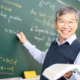 High school calculus survey: Older Asian male with gray hair and glasses in white business shirt with navy vest stands at chalkboard writing calculus formulas while smiling towards students