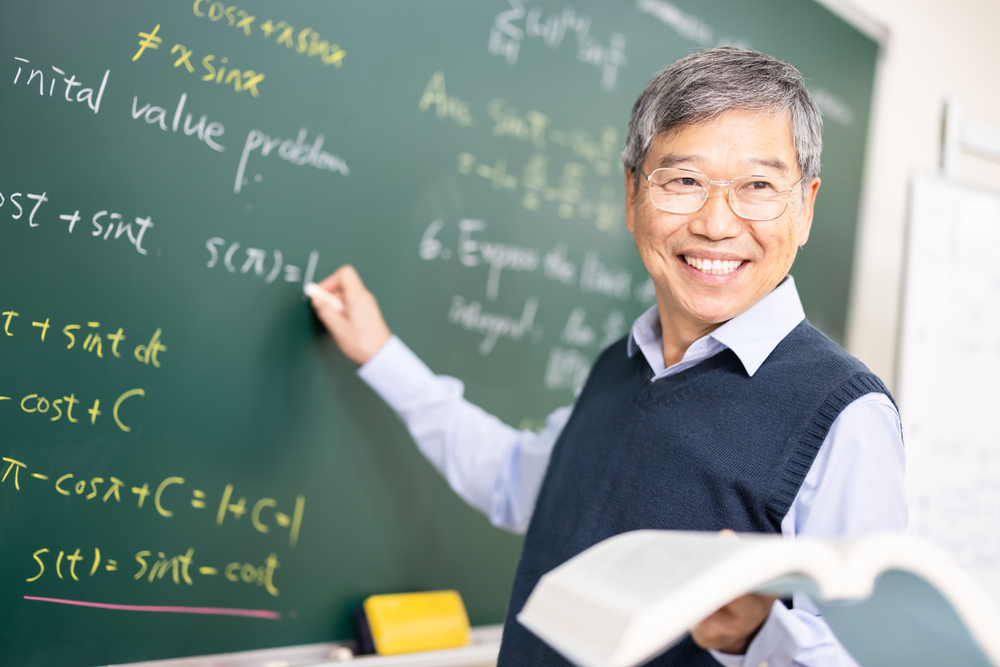 High school calculus survey: Older Asian male with gray hair and glasses in white business shirt with navy vest stands at chalkboard writing calculus formulas while smiling towards students