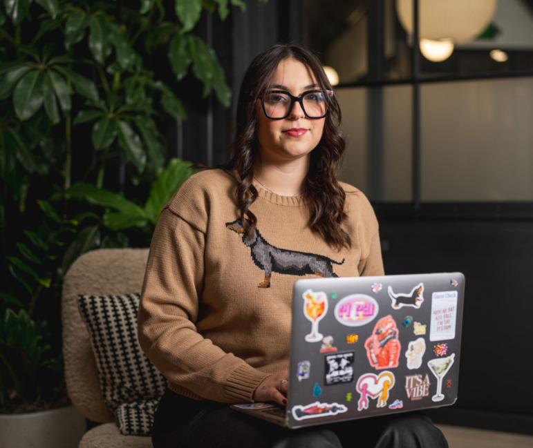 AI in colleges: Young woman with long dark hair and dark-framed glasses and tan long-sleeved top, sit in front of greenery with laptop in her lap, the back of laptop screen covered in stickers