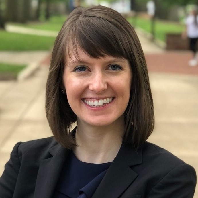 AI in College: Woman with shoulder length dark hair in black suit and top stands on outdoor sidewalk lined with trees and lawns.