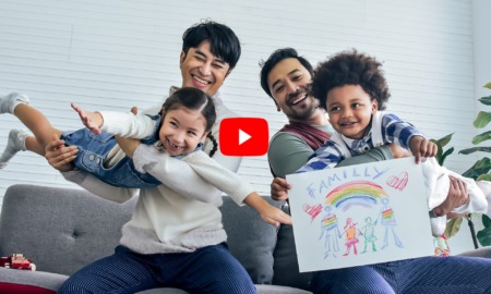 LGBTQ adoptions: Male gay couple sitting on sofa in living room holding two happy toddlers -- Caucasian girl and Black boy -playing with crayon drawing of rainbow with text "Family".