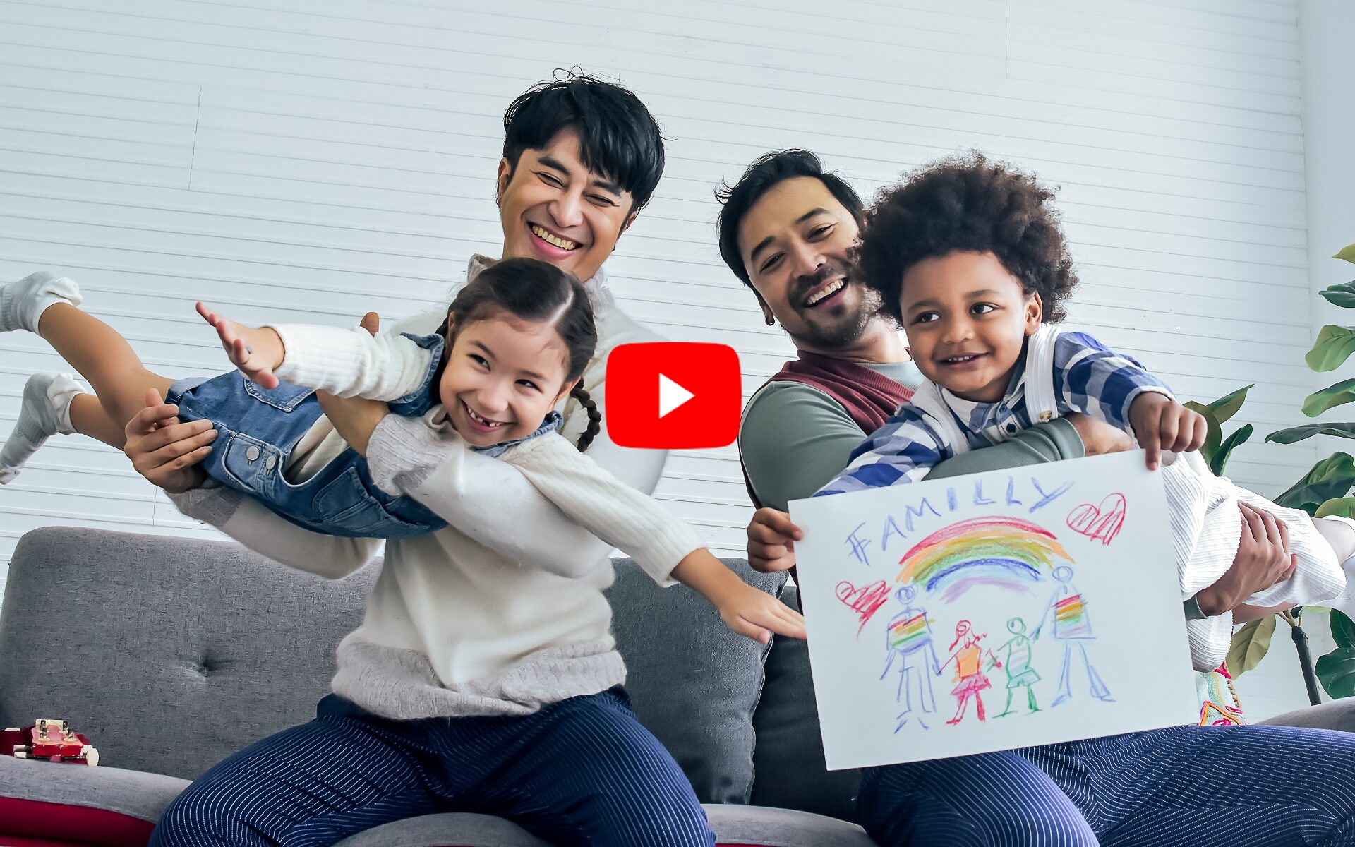 LGBTQ adoptions: Male gay couple sitting on sofa in living room holding two happy toddlers -- Caucasian girl and Black boy -playing with crayon drawing of rainbow with text "Family".