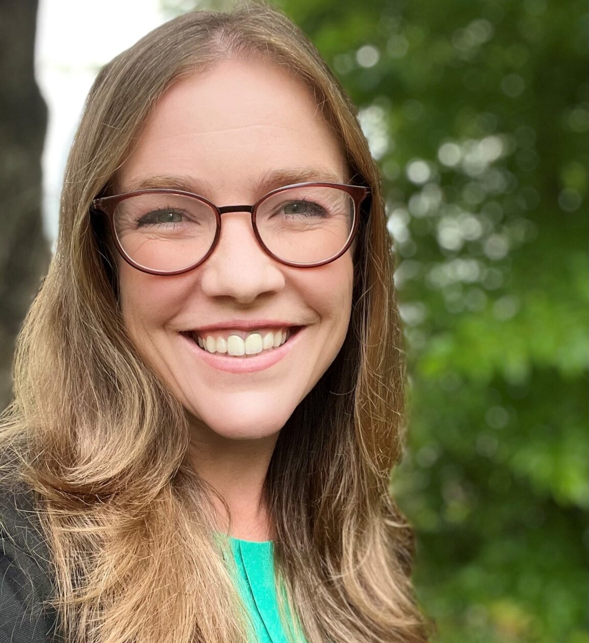 LGBTQ adoptions: Headshot Meg York -- young woman with long blonde hair and dark-framed glasses in teal top and dark jacket smiles into camera with greenery in the background.