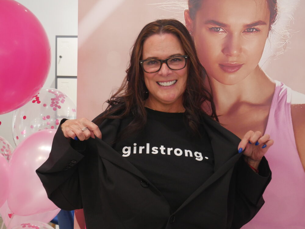 Girlstrong fashion: Young woman with long dark hair and dark-framed glasses wears black t-shirt with "Girlstrong" in white letters on chest