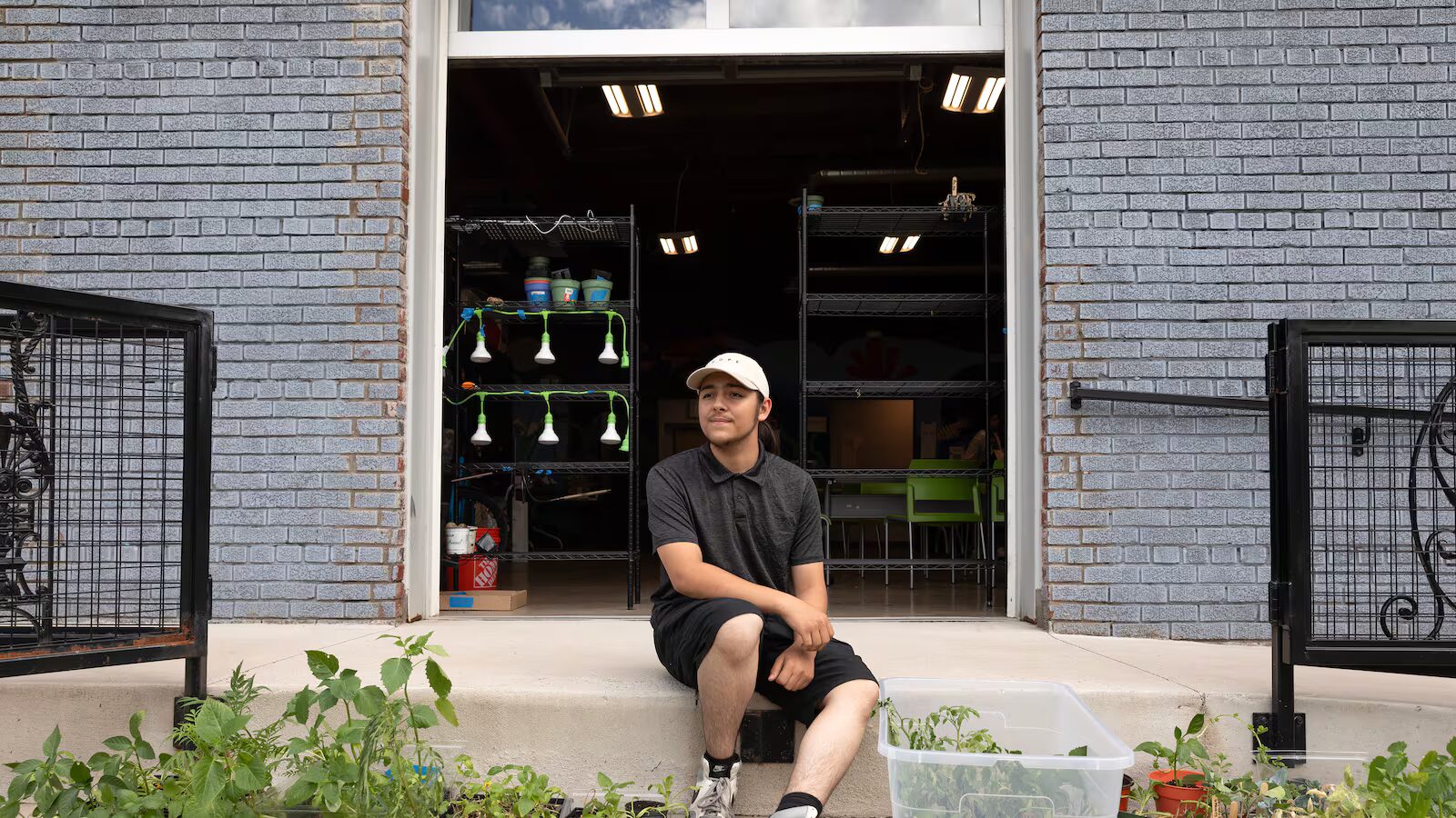 High school drop-outs: Young man with long dark hair pulled back in light baseball cap, black shirt & shorts, sits on wide cement steps with black railings in front of large open roll-up doorway into dark shop, interior