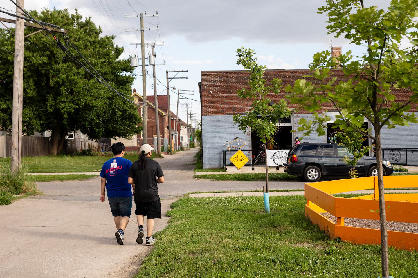 High school drop-outs: Two adults in black clothes walk with arms around each other's shlulders on a grass-lined sidewalk towards a two-story red and white building about a block way