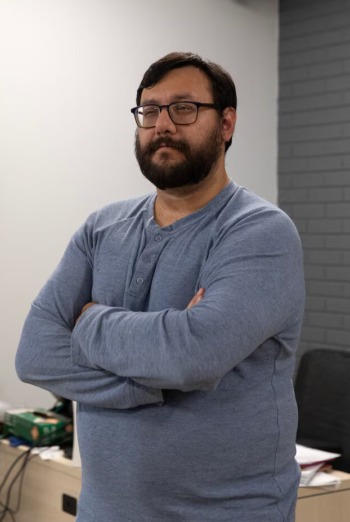 High school drop-out: Man with short brown hir and full beard wearing light blue sweater stands in front of white wall with arms crossed