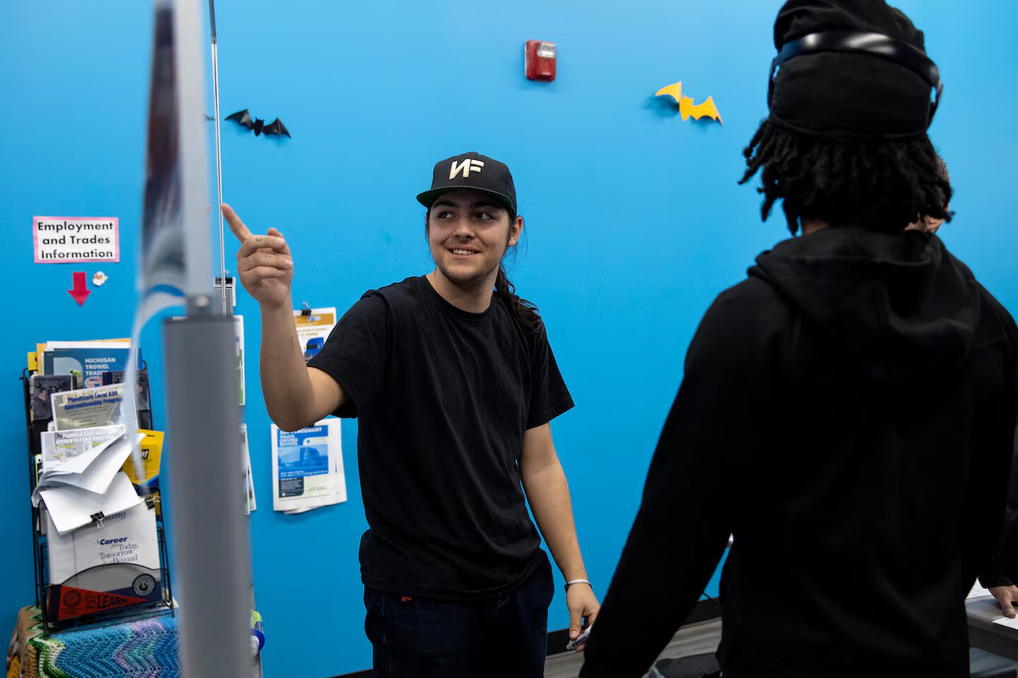 High school drop-out: Young man with long dark hair pulled back in black baseball cap, black shirt ab black pants stands in front of bright turquoise wall pointing right hand while speaking to person in all black with back turned to camera