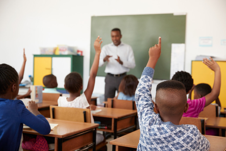 Black teachers leaving Philadelphia: Black man in white shirt and brown pants stands in front of green chalkboard facing several Black elementary students sitting at desks with hands raised