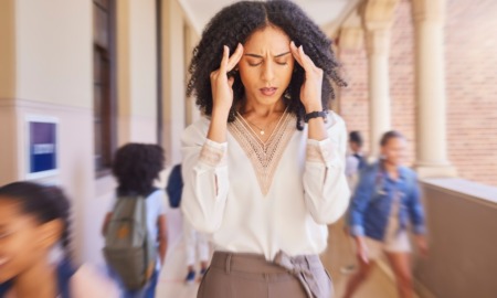 Black teachers leaving Philadelphia: Black woman in white blouse and tan skirt standing in hallway full of children holding head in her hands with eyes closed and grimacing face