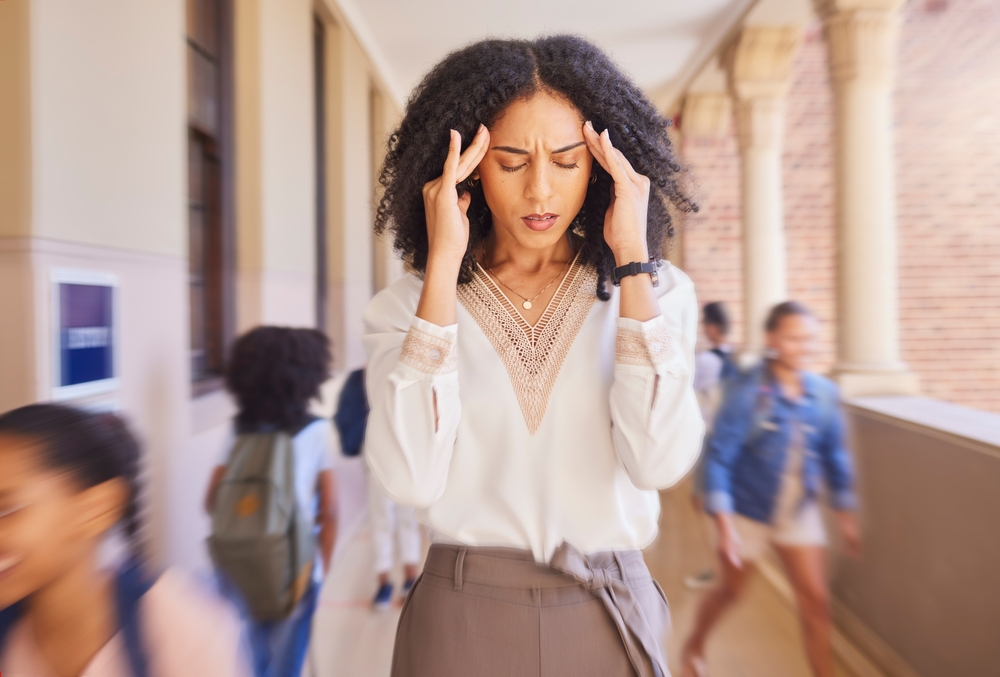 Black teachers leaving Philadelphia: Black woman in white blouse and tan skirt standing in hallway full of children holding head in her hands with eyes closed and grimacing face