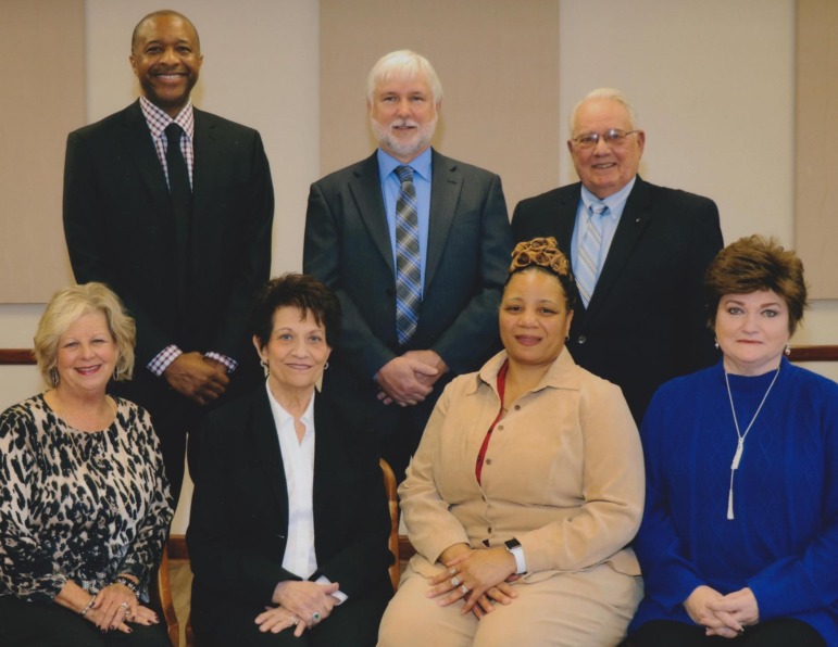 Amite school segregation academies: Four middle age women (3 white; one Black) are seated with hands folded in laps in front of three middle age males (2 white, one Black) standing with hands clasped. All are wearing business attire.