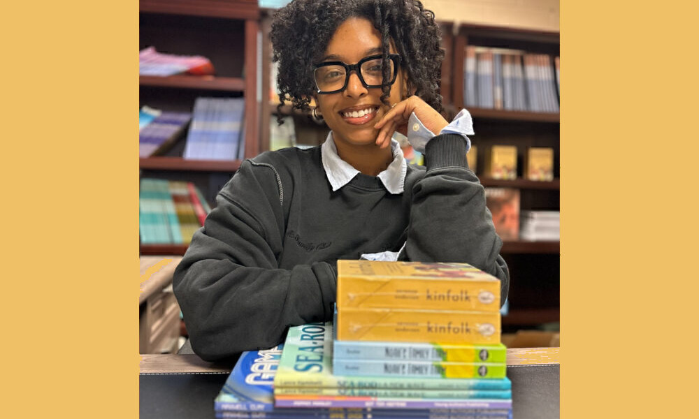 Youth publisher: Young Black woman with short black hair and black-rimmed glasses in dark gray sweatshirt wits at table with stck of books and bookshelves in background.