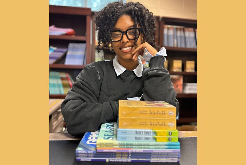 Youth publisher: Young Black woman with short black hair and black-rimmed glasses in dark gray sweatshirt wits at table with stck of books and bookshelves in background.