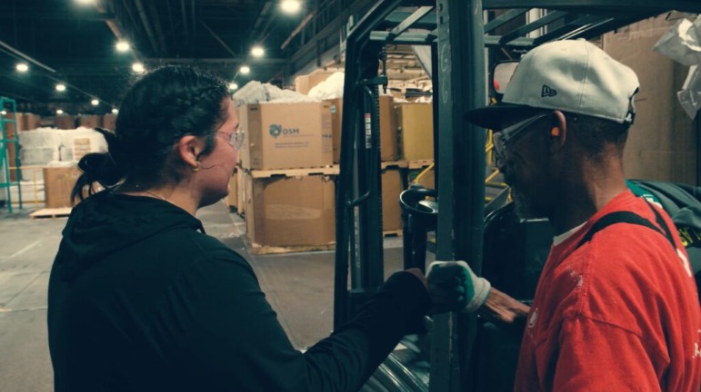 Apprenticeships Indiana: A young adult woman with dark hair pulled back stands with an older Black man in hardhat speaking to each other while doing a fist bump on the floor of an industrial warehouse full of containers and equipment.