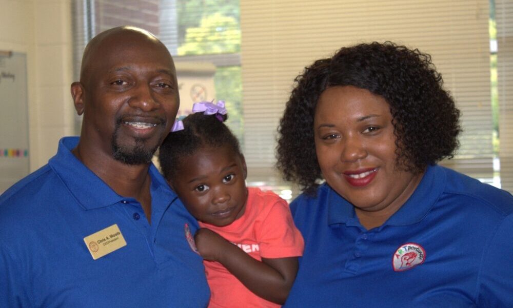 Nonprofit children's art program: A Black man with black beard and Black women wit bobbed black hair both in bright blue polo shirts hold a toddle wearing a coral top