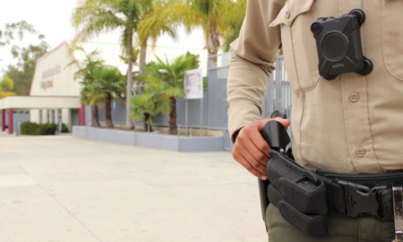 Police in schools: Adult male in tan shirt and brown pants, close-up of chest with body cam and holster with gun, with male hand on gun butt, and a school campus entrnce in the background.