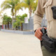 Police in schools: Adult male in tan shirt and brown pants, close-up of chest with body cam and holster with gun, with male hand on gun butt, and a school campus entrnce in the background.