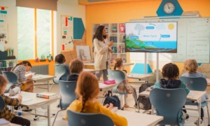 Curriculum fidelity k-12: Woman with long, dark hair in tan sweater and slacks stands at front of colorful classroom next to slide screen with a pointer explaining a colorful science slide to elementary students sitting at desks